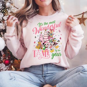 May include: Pink sweatshirt with a Christmas-themed design featuring Mickey Mouse, Minnie Mouse, Donald Duck, Goofy, and Pluto. The text on the sweatshirt reads "It's the most Wonderful Time of the Year".