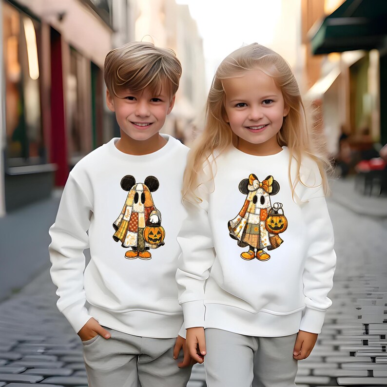 May include: Two children wearing white sweatshirts with Halloween-themed graphics. The graphics feature a ghost character with Mickey Mouse ears, a patchwork design, and a jack-o'-lantern. The children are wearing gray pants.