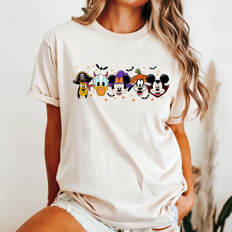 Op de afbeelding: Een beige T-shirt met een grafische print van Mickey Mouse, Donald Duck, Goofy en Pluto in Halloween-kostuums. De personages staan in een rij met vleermuizen en oranje stippen op de achtergrond.