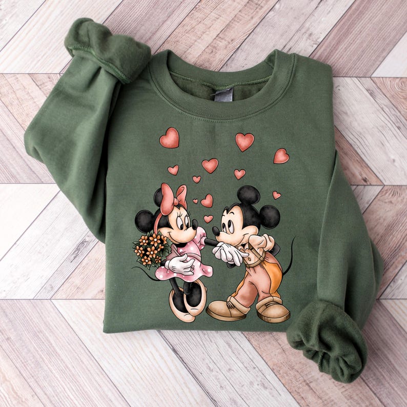 May include: A green sweatshirt with a graphic print of Mickey Mouse and Minnie Mouse holding hands.  Pink hearts are floating around them.