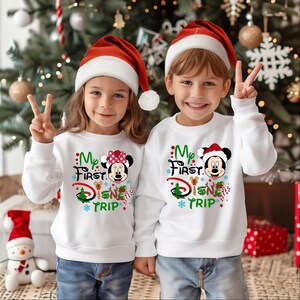 May include: Two children wearing white sweatshirts with a Christmas-themed design featuring Mickey Mouse and the text "My First Disney Trip". The children are wearing red Santa hats and smiling.