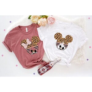 May include: Two t-shirts with a leopard print Mickey Mouse design. One shirt is a dusty rose color and the other is white.