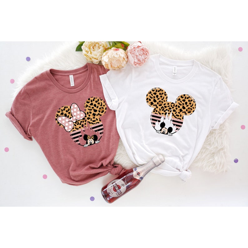 May include: Two t-shirts with a leopard print Mickey Mouse design. One shirt is a dusty rose color and the other is white.
