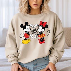Może przedstawiać: Beżowa bluza z nadrukiem przedstawiającym Myszki Miki i Minnie trzymające się za ręce w kształcie serca. Tekst "sweet love" jest nadrukowany czarnym kolorem pod postaciami.