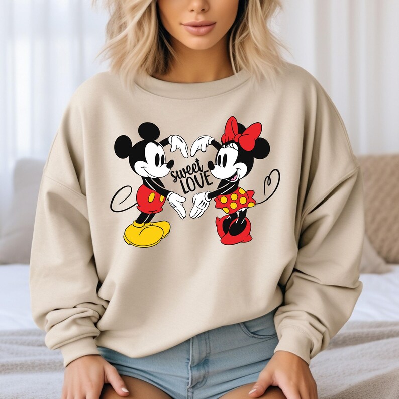 Może przedstawiać: Beżowa bluza z nadrukiem przedstawiającym Myszki Miki i Minnie trzymające się za ręce w kształcie serca. Tekst "sweet love" jest nadrukowany czarnym kolorem pod postaciami.