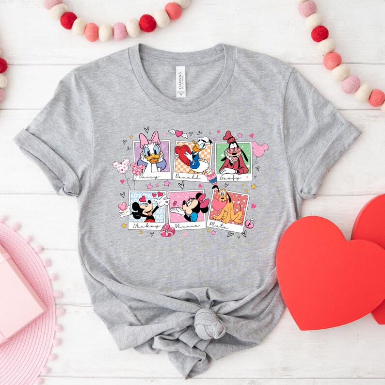 K&ouml;nnte beinhalten: Ein graues T-Shirt mit einem grafischen Druck von Disney-Charakteren in Polaroid-Rahmen. Die Charaktere sind Mickey Mouse, Minnie Mouse, Pluto, Donald Duck, Daisy Duck und Goofy. Der Text auf dem Shirt lautet "Mickey", "Minnie", "Pluto", "Daisy", "Donald" und "Goofy".