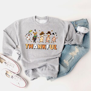 May include: A light gray sweatshirt with a graphic featuring cartoon dogs in fall attire. The word "THANKFUL" is printed in orange and white. The sweatshirt is paired with blue jeans and white sneakers.