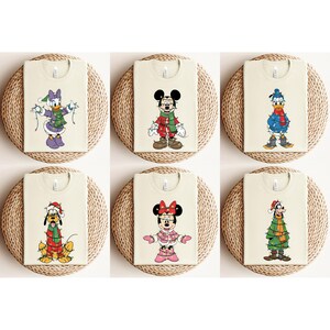 May include: Six cream-colored t-shirts featuring Disney characters in festive holiday attire. Characters include Mickey Mouse, Minnie Mouse, Donald Duck, Daisy Duck, Pluto, and Goofy, each adorned with Christmas lights and seasonal clothing.