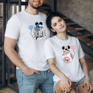 May include: Two light gray t-shirts featuring cartoon robot designs with Mickey Mouse ears and heart-shaped balloons. One robot is blue and white, the other is pink and white. The shirts are worn by a man and a woman.