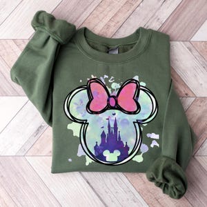 May include: Green sweatshirt with a watercolor-style Mickey Mouse head design. The design features a pink bow and a silhouette of Cinderella's castle inside the ears.