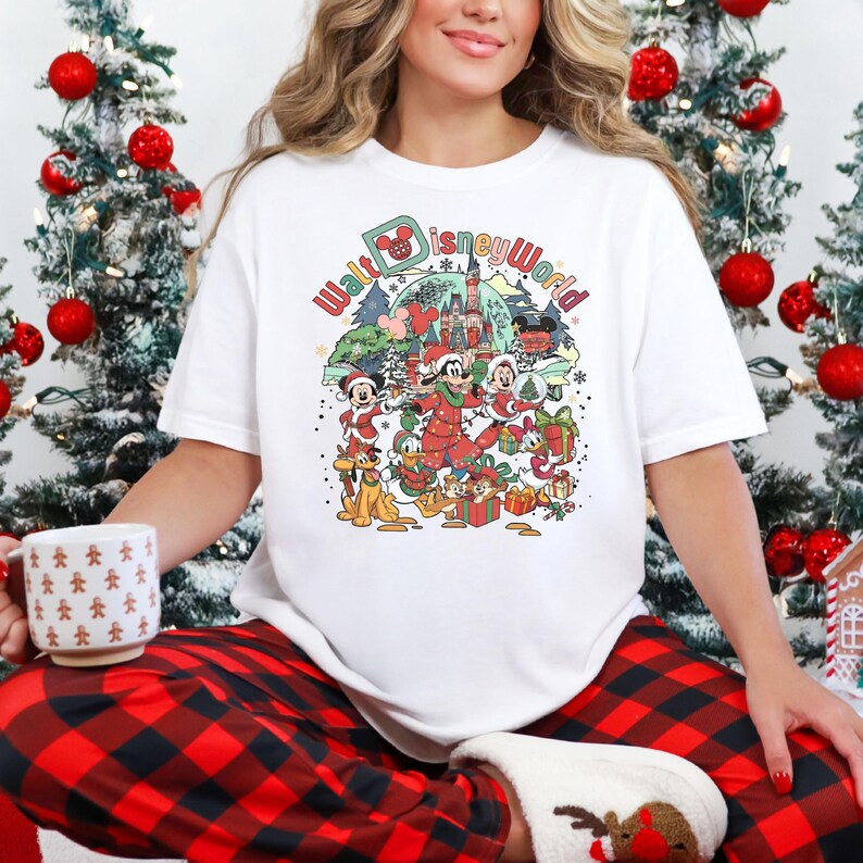 May include: White t-shirt featuring a colorful graphic of Disney characters celebrating Christmas at Walt Disney World. The design includes Mickey Mouse, Donald Duck, and Goofy, with the park's castle in the background. The text "Walt Disney World" is also included.