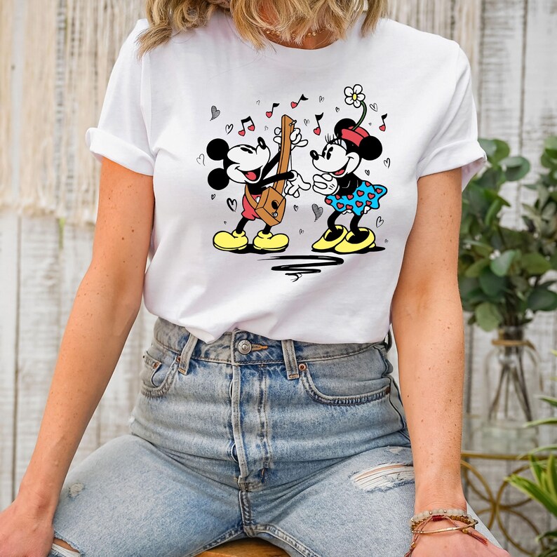 Pode incluir: Camiseta branca com um gr&aacute;fico de Mickey Mouse e Minnie Mouse tocando m&uacute;sica. Mickey est&aacute; tocando guitarra e Minnie est&aacute; cantando. H&aacute; cora&ccedil;&otilde;es e notas musicais ao redor deles.