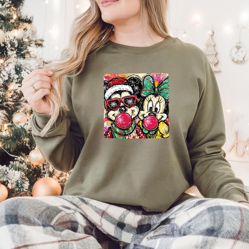 May include: An olive green sweatshirt featuring a colorful graphic of Mickey and Minnie Mouse. Mickey wears a Santa hat and sunglasses, while Minnie has a bow. Both characters are blowing bubblegum bubbles. The sweatshirt is worn by a person.