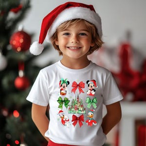 May include: A white t-shirt with a Christmas theme featuring Mickey Mouse, Minnie Mouse, Donald Duck, and a Christmas castle. The shirt has red and green bows and a red Santa hat.