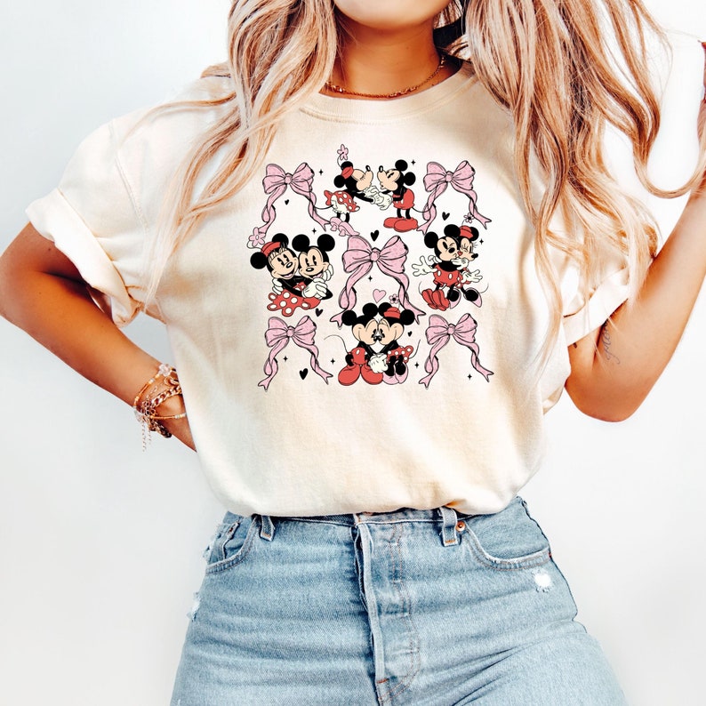 May include: A white t-shirt with a pattern of Mickey Mouse and Minnie Mouse in various poses. The pattern includes pink bows and hearts.
