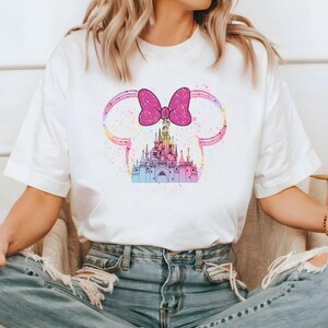 May include: White t-shirt with a pink bow and a colorful castle graphic. The castle is surrounded by a pink and white outline resembling Mickey Mouse ears.