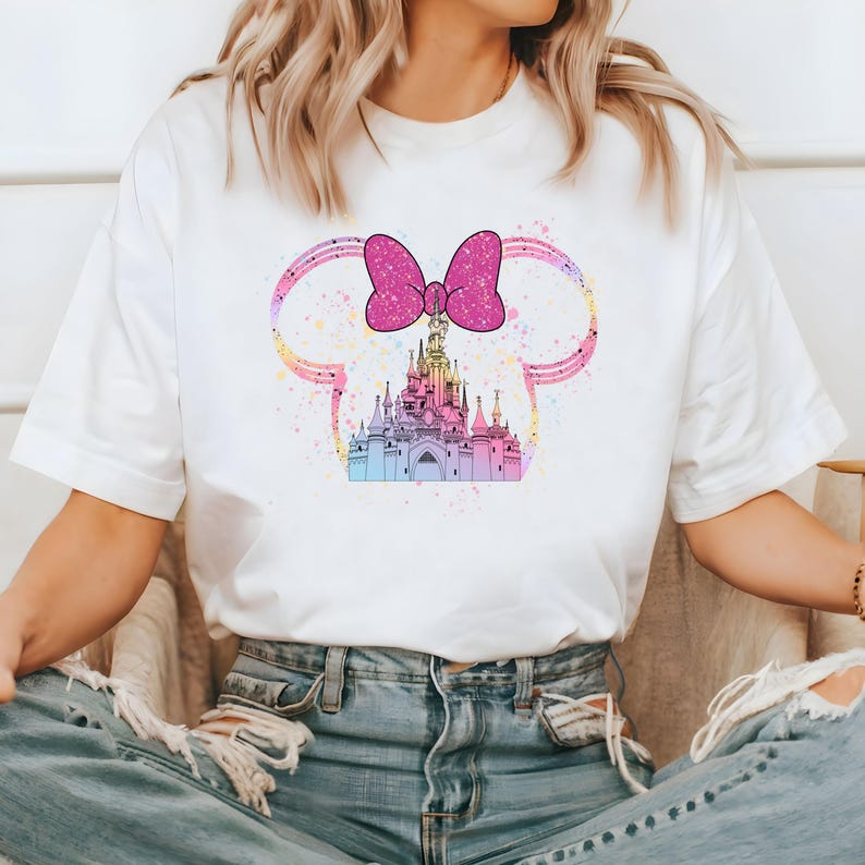 May include: White t-shirt with a pink bow and a colorful castle graphic. The castle is surrounded by a pink and white outline resembling Mickey Mouse ears.