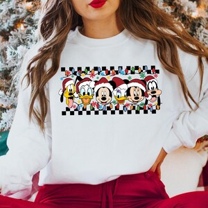 May include: White sweatshirt with a checkered pattern and a row of Disney characters wearing Santa hats. The characters include Mickey Mouse, Minnie Mouse, Donald Duck, Daisy Duck, and Pluto. The characters are surrounded by Christmas lights and snowflakes.