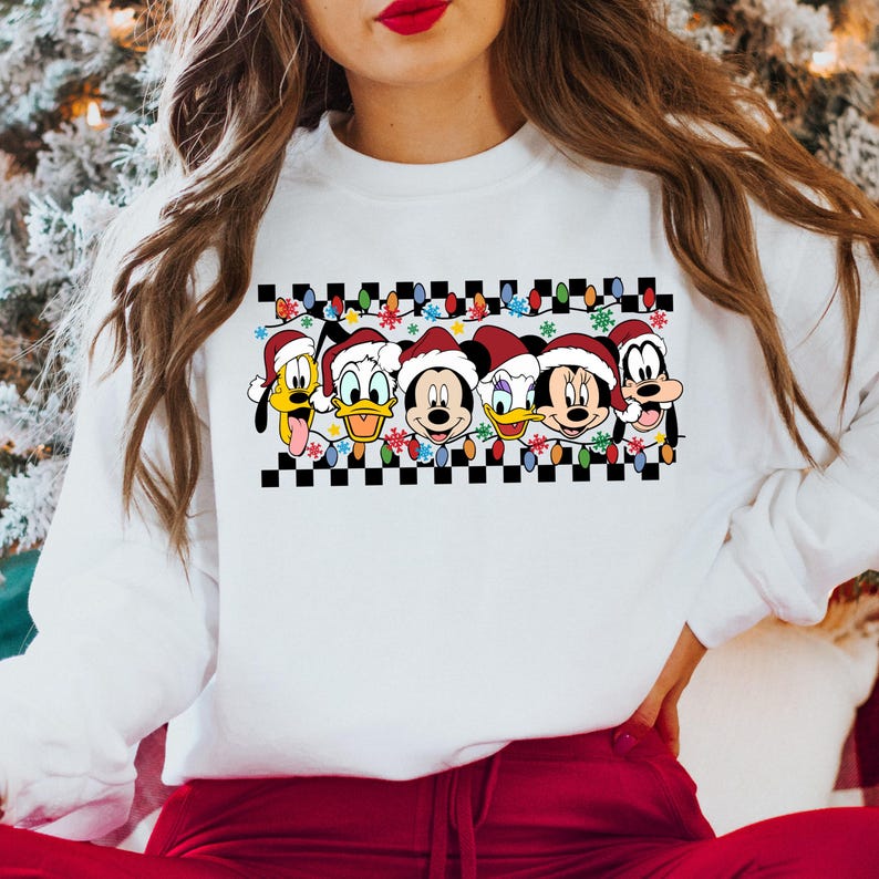 May include: White sweatshirt with a checkered pattern and a row of Disney characters wearing Santa hats. The characters include Mickey Mouse, Minnie Mouse, Donald Duck, Daisy Duck, and Pluto. The characters are surrounded by Christmas lights and snowflakes.
