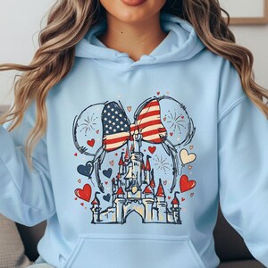 May include: Light blue hoodie featuring a graphic of Mickey Mouse ears with an American flag design. The design includes a castle, hearts, and fireworks. The hoodie has a front pocket and a drawstring hood.