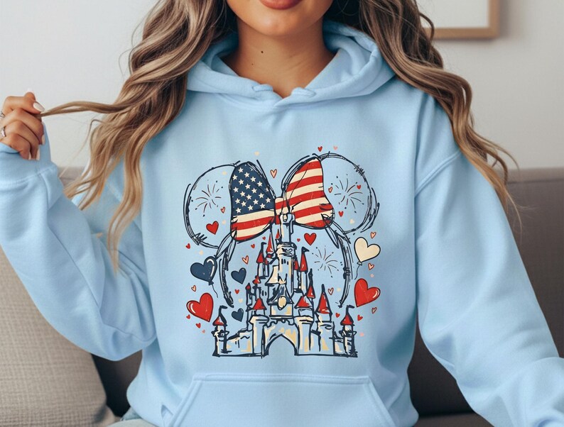 May include: Light blue hoodie featuring a graphic of Mickey Mouse ears with an American flag design. The design includes a castle, hearts, and fireworks. The hoodie has a front pocket and a drawstring hood.