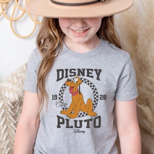 May include: A grey t-shirt with a vintage-style graphic of Pluto, the Disney dog. The graphic features Pluto in a circle with a checkered border and the text "DISNEY 19 PLUTO 28".