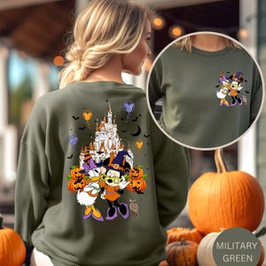 May include: Military green sweatshirt featuring a Halloween-themed graphic of Minnie Mouse and Daisy Duck in witch costumes, with a castle, pumpkins, and bats. The sweatshirt has a crew neck and long sleeves. The color is labeled "Military Green".