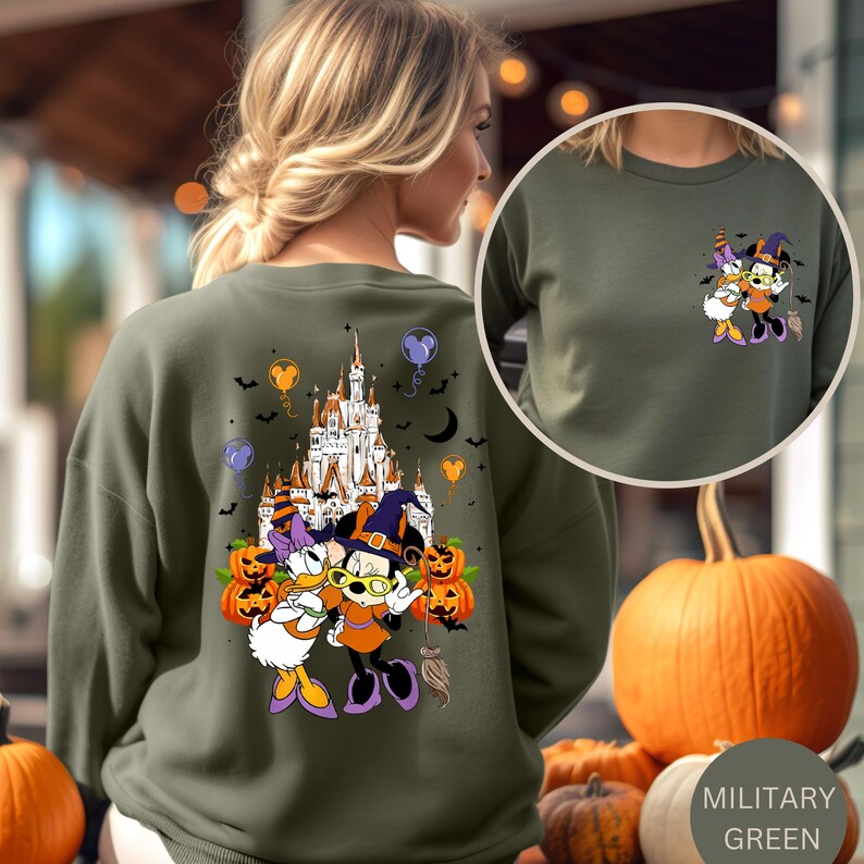 May include: Military green sweatshirt featuring a Halloween-themed graphic of Minnie Mouse and Daisy Duck in witch costumes, with a castle, pumpkins, and bats. The sweatshirt has a crew neck and long sleeves. The color is labeled "Military Green".