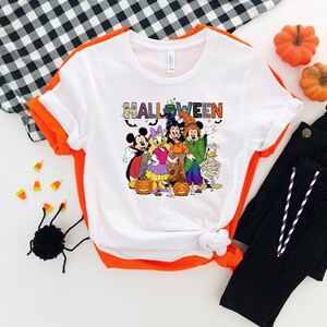 May include: White t-shirt with orange trim featuring a Halloween design. The design includes the word "HALLOWEEN" in colorful letters and cartoon characters dressed in Halloween costumes. Black pants, pumpkins, and candy corn are also visible.