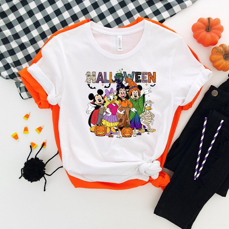 May include: White t-shirt with orange trim featuring a Halloween design. The design includes the word "HALLOWEEN" in colorful letters and cartoon characters dressed in Halloween costumes. Black pants, pumpkins, and candy corn are also visible.