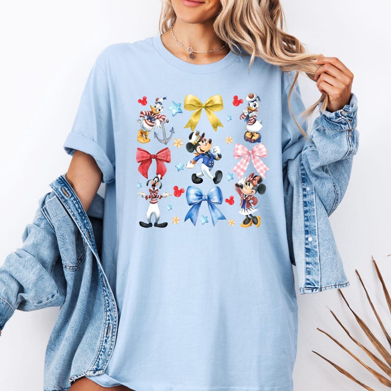 May include: Light blue t-shirt with a colorful print of Mickey Mouse, Minnie Mouse, Donald Duck, and Goofy wearing sailor outfits and surrounded by red, blue, and yellow bows.