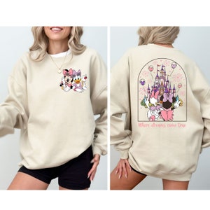Pu&ograve; includere: Una felpa beige con un'immagine di cartone animato di Topolino e Paperina sul davanti e un'immagine di cartone animato del castello di Cenerentola con Topolino e Paperina sul retro. Il testo "Where dreams come true" &egrave; stampato sotto l'immagine sul retro.