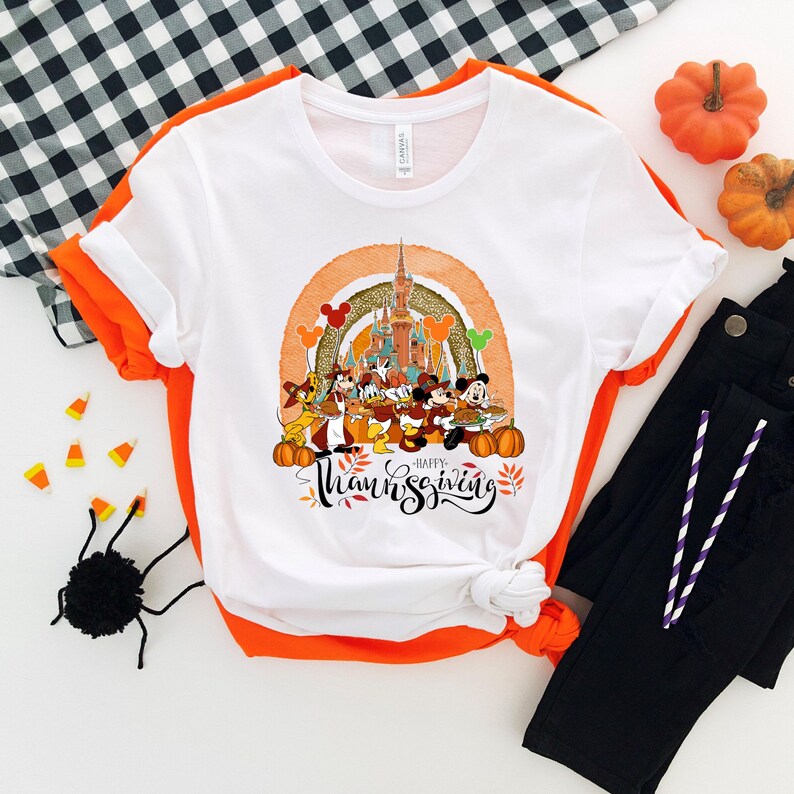 May include: White t-shirt with a colorful graphic featuring Disney characters, a castle, and the text "Happy Thanksgiving." The design includes pumpkins, Mickey Mouse balloons, and a rainbow arch. The shirt is paired with black pants and orange accents.