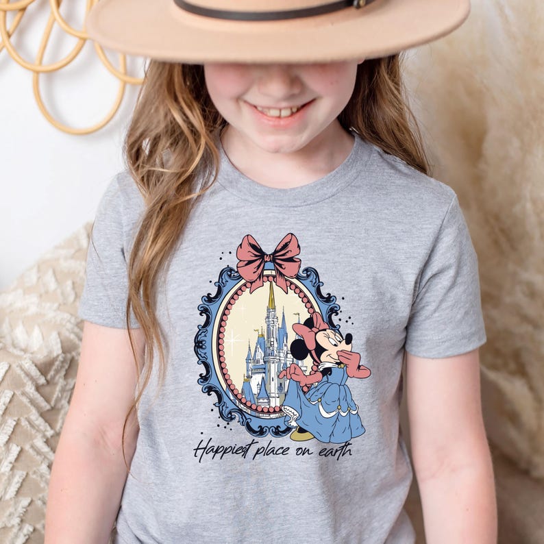 May include: A grey t-shirt with a vintage-style illustration of Mickey Mouse in front of Cinderella's Castle. The illustration is framed with a blue and gold ornate frame and a pink bow. The text "Happiest place on earth" is printed below the illustration.