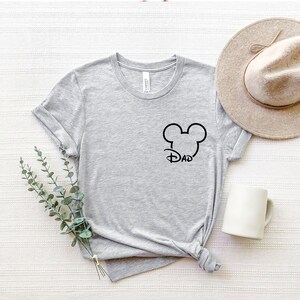 Puede incluir: Camiseta gris con un dise&ntilde;o de cabeza de Mickey Mouse negro y la palabra "DAD" dentro de las orejas.