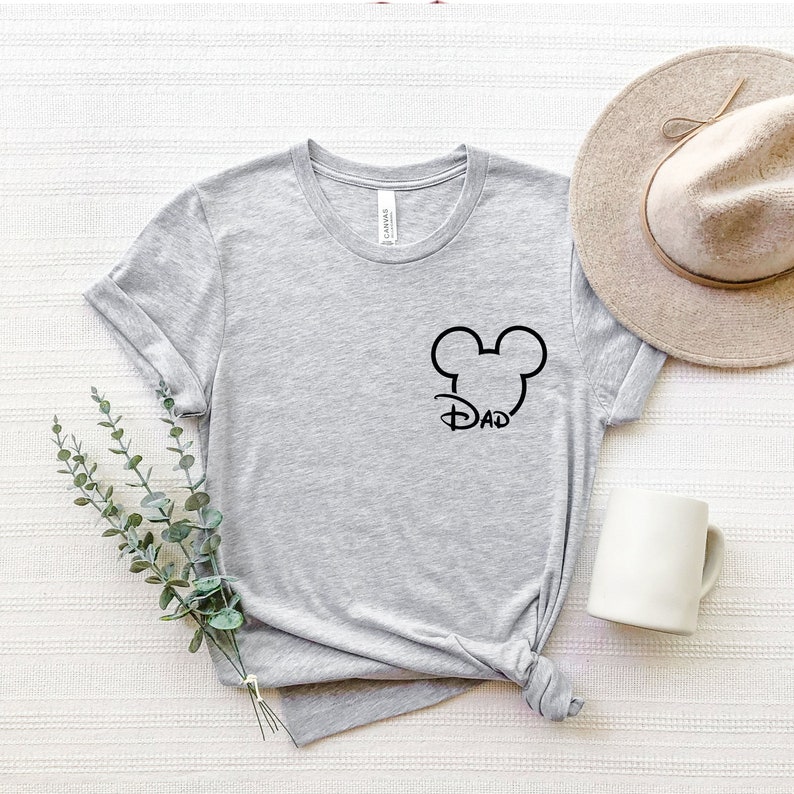 Puede incluir: Camiseta gris con un dise&ntilde;o de cabeza de Mickey Mouse negro y la palabra "DAD" dentro de las orejas.