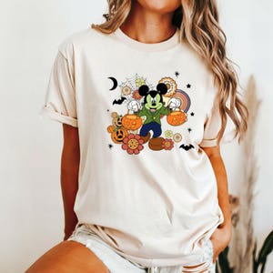 K&ouml;nnte beinhalten: Beiges T-Shirt mit einem Retro-Halloween-Design mit Micky Maus. Micky tr&auml;gt eine gr&uuml;ne Jacke und blaue Hose und h&auml;lt zwei K&uuml;rbis-Eimer. Das Design umfasst Blumen, Flederm&auml;use, Sterne und einen Regenbogen.