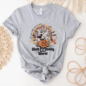 Puede incluir: Camiseta gris claro con un gr&aacute;fico de Halloween de personajes de Disney, incluyendo Mickey Mouse, Donald Duck y Goofy, frente a un castillo y una calabaza. El texto "Walt Disney World" est&aacute; debajo.