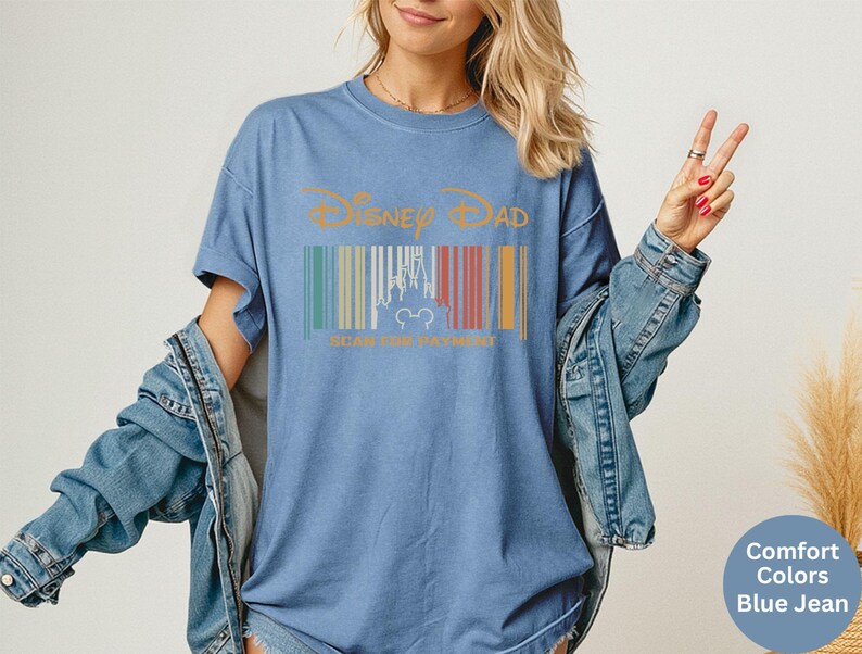 May include: Blue jean-coloured t-shirt with the text "Disney Dad" in gold. The shirt features a barcode design with a castle silhouette and Mickey Mouse ears. The shirt is a comfort colour.