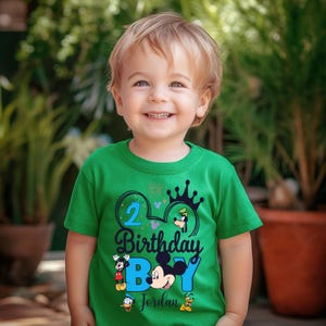 Op de afbeelding: Een groen T-shirt met een blauw en zwart Mickey Mouse-ontwerp. Het shirt heeft de tekst "2 Birthday Boy Jordan" en bevat andere Disney-figuren zoals Goofy, Donald Duck en Pluto.