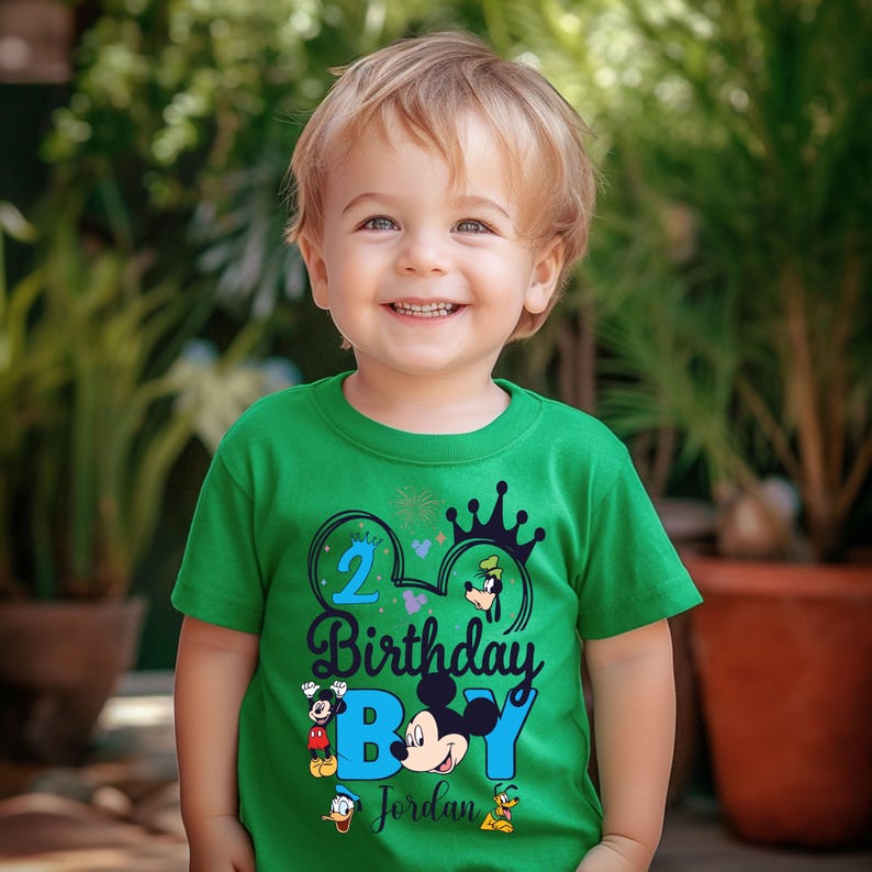 Op de afbeelding: Een groen T-shirt met een blauw en zwart Mickey Mouse-ontwerp. Het shirt heeft de tekst "2 Birthday Boy Jordan" en bevat andere Disney-figuren zoals Goofy, Donald Duck en Pluto.