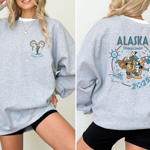May include: A heather gray sweatshirt with a crew neck. The front features a small graphic of Mickey Mouse and the words "Alaska Disney Cruise". The back displays a larger design with Disney characters, the words "Alaska Disney Wonder" and the year 2025.