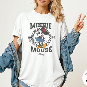 May include: White t-shirt featuring a distressed graphic of Minnie Mouse with the text "MINNIE MOUSE 1928". The design includes a checkered circle and the Disney logo. The shirt is paired with a denim jacket.