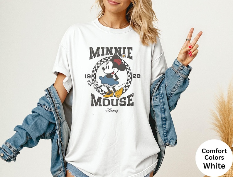 May include: White t-shirt featuring a distressed graphic of Minnie Mouse with the text "MINNIE MOUSE 1928". The design includes a checkered circle and the Disney logo. The shirt is paired with a denim jacket.