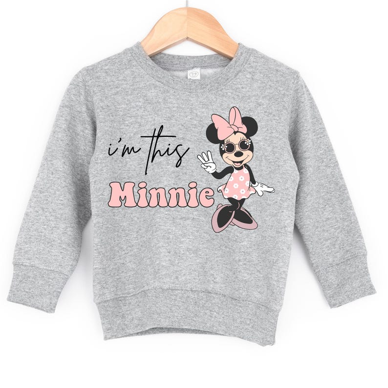 Puede incluir: Sudadera gris con un gr&aacute;fico de Minnie Mouse rosa y negro y el texto "i'm this Minnie" en negro.