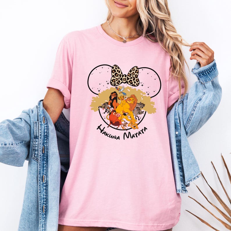 Puede incluir: Camiseta rosa con un dise&ntilde;o de orejas de Mickey Mouse en blanco y negro con los personajes de El Rey Le&oacute;n y el texto "Hakuna Matata".