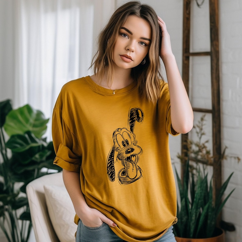 May include: Mustard yellow t-shirt featuring a black sketch of a cartoon dog with its tongue out. The shirt has rolled-up sleeves and a relaxed fit. The model is wearing the shirt. The background includes plants and a wooden ladder.
