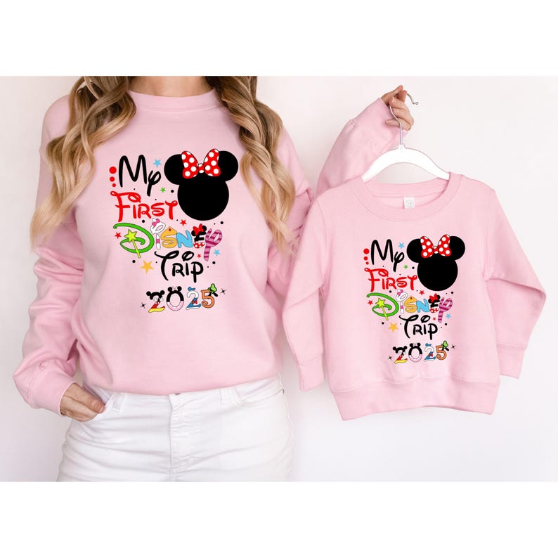 Peut inclure: Deux sweat-shirts roses avec l'inscription "My First Trip 2025" et un motif Minnie Mouse. L'un est port&eacute;, l'autre est sur un cintre. Le motif Minnie Mouse comprend un n&oelig;ud rouge &agrave; pois blancs.