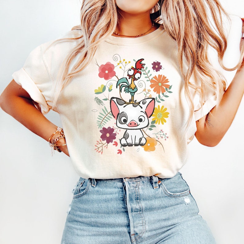 Puede incluir: Una camiseta blanca con una imagen de dibujos animados de un cerdo y un gallo en un dise&ntilde;o floral. El cerdo es blanco con manchas negras y el gallo es rojo, amarillo y verde. Las flores son rosas, amarillas, naranjas y moradas.