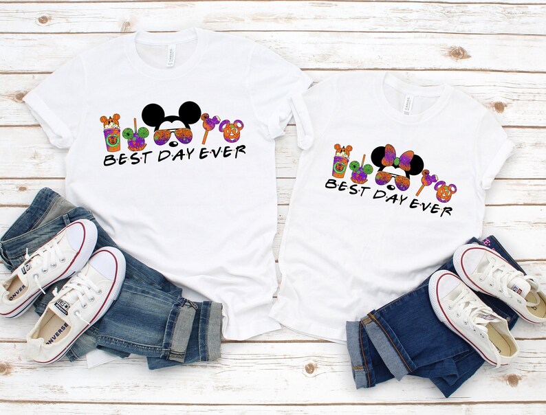 May include: Two white t-shirts with a Mickey Mouse and Minnie Mouse design. The shirts have the text "Best Day Ever" and feature a variety of Halloween-themed icons.